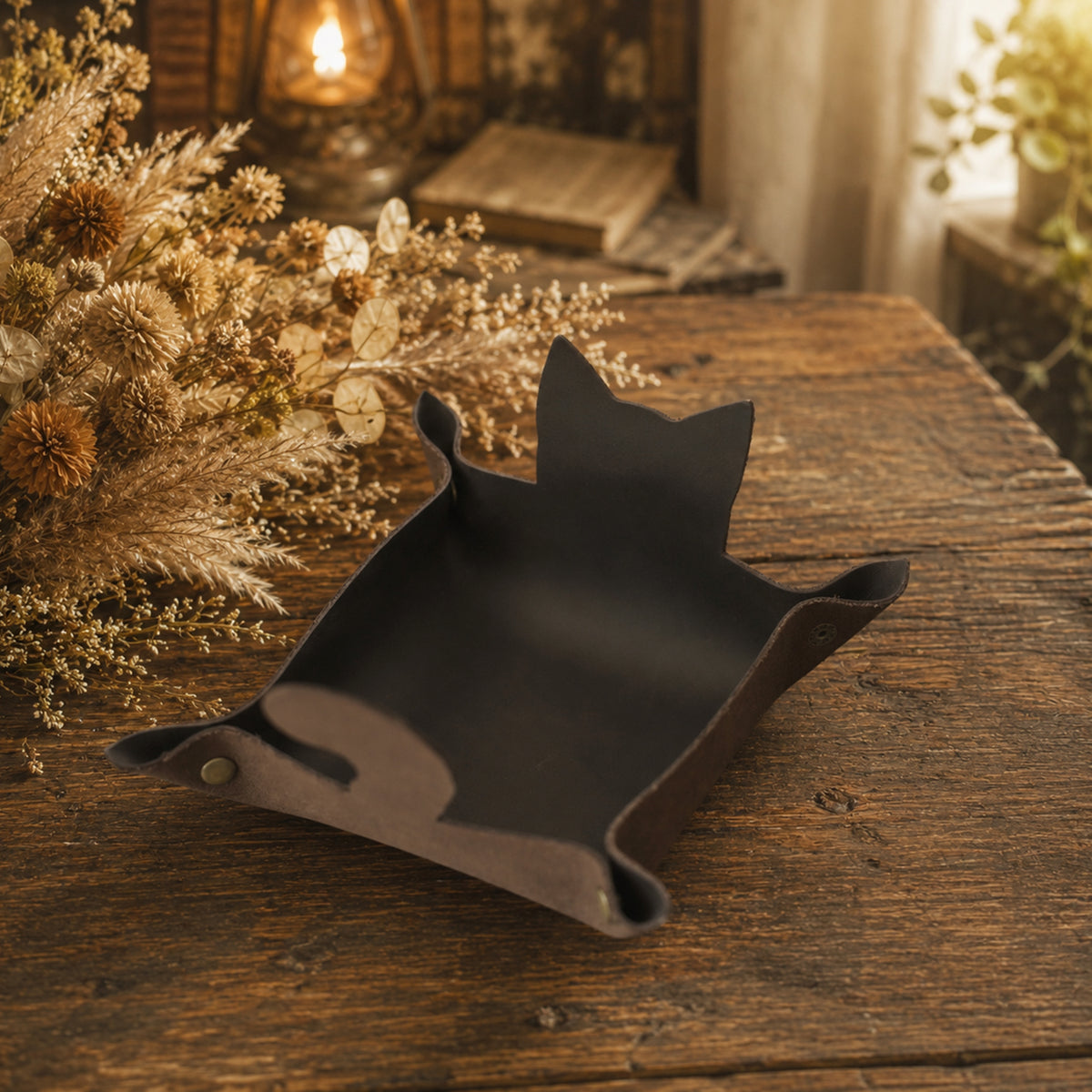 A dark brown leather cat-shaped tray with a small metal snap button, resting on a rustic wooden table with dried flowers and a warm lantern in the background.