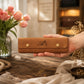 A hand holds a tan leather jewelry organizer case with gold snap buttons and an engraved monogram, set on a rustic wooden table with pink tulips and jewelry in a warm, softly lit interior.