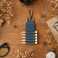 A dark blue leather golf club holder with a brass clasp and five pegs, displayed on a rustic wooden table surrounded by dried flowers, a camera lens, glasses, a pocket watch, and a vintage card.