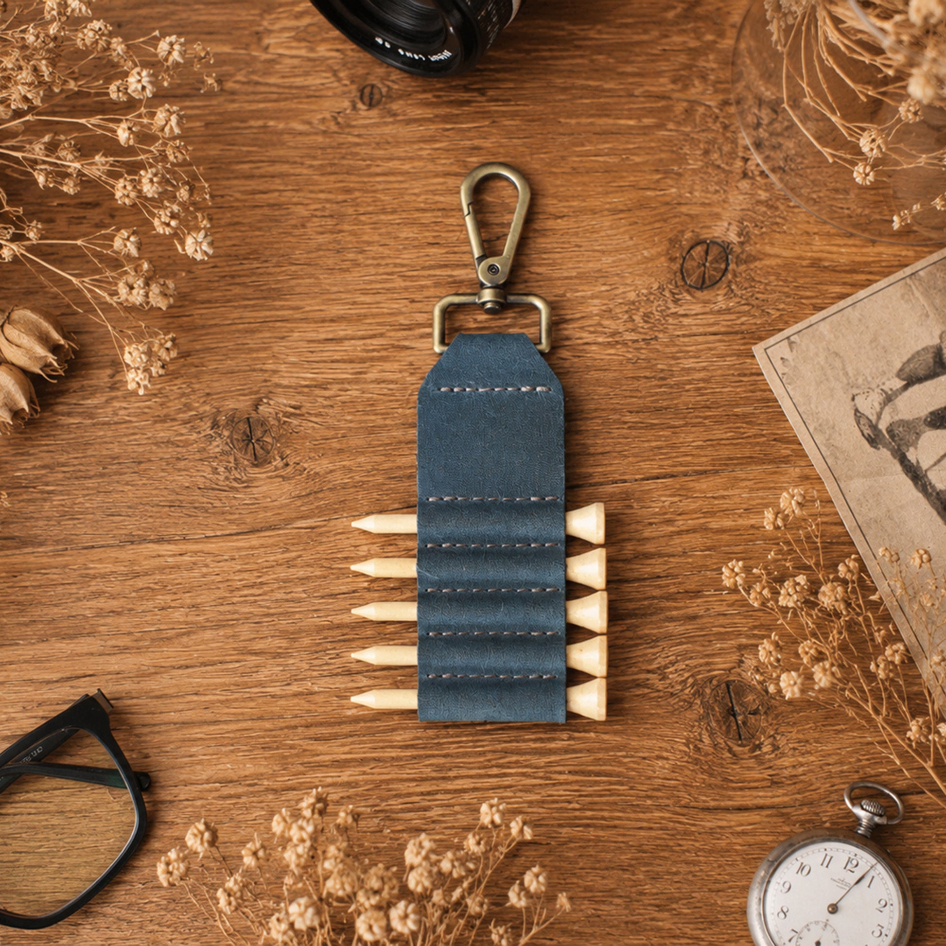 A dark blue leather golf club holder with a brass clasp and five pegs, displayed on a rustic wooden table surrounded by dried flowers, a camera lens, glasses, a pocket watch, and a vintage card.