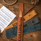 Three personalized drumstick pouches in brown, tan, and slate blue, laid out on a rustic wooden table with a sheet of music and rope props.