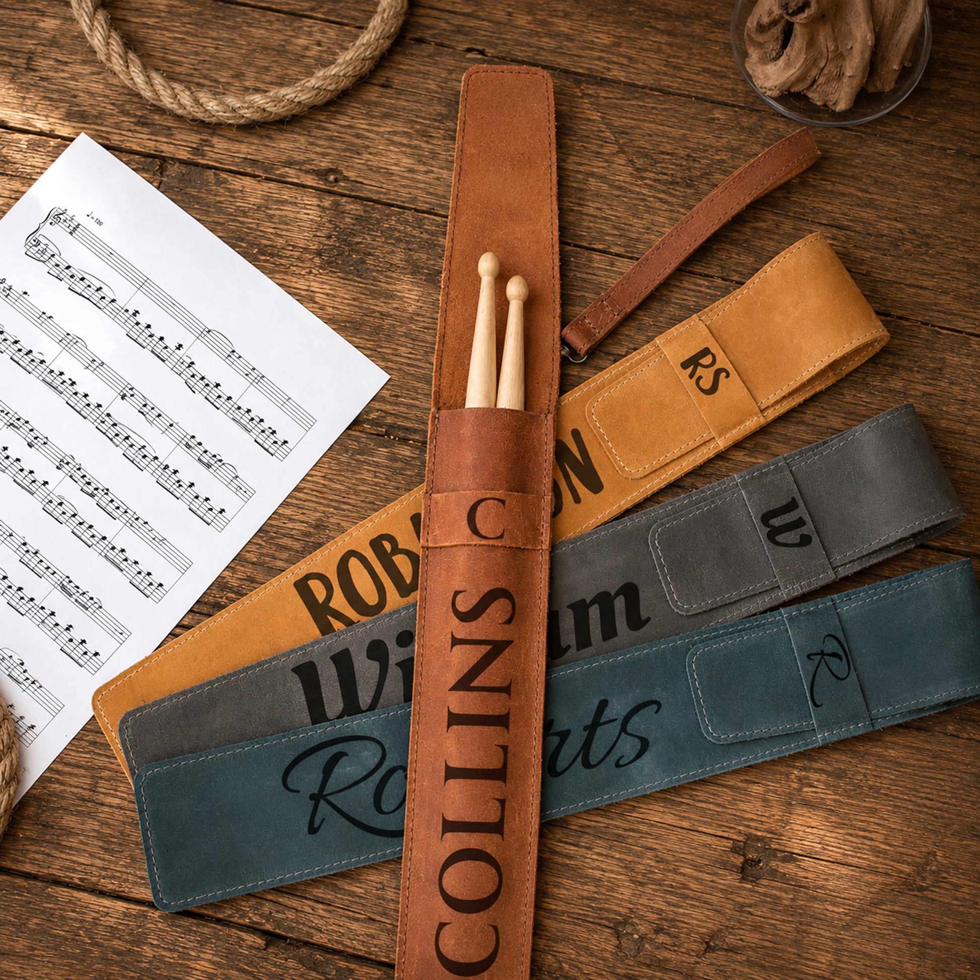 Three personalized drumstick pouches in brown, tan, and slate blue, laid out on a rustic wooden table with a sheet of music and rope props.