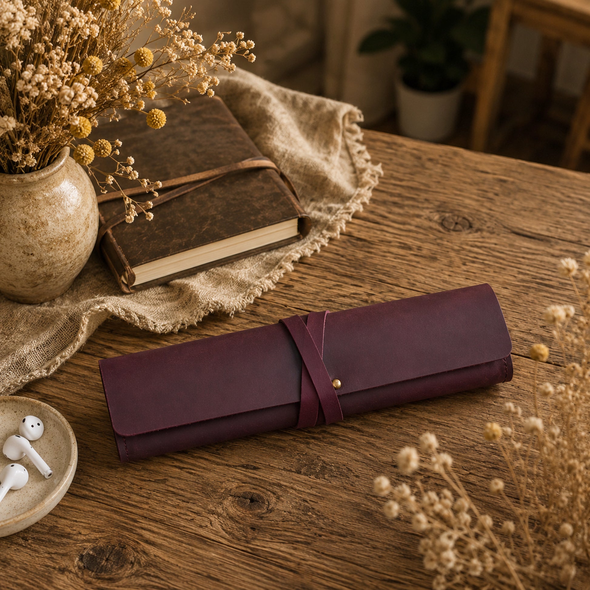 A rolled burgundy leather journal or notebook cover with a wrap strap and gold-tone button, resting on a rustic wooden table. The scene is styled with a vintage book, a vase of dried flowers, white earbuds, and a burlap cloth.