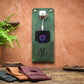 A green leather wall-mounted charging stand with a front pocket holding a smartphone, shown in a styled interior setting against a terracotta wall. Two additional charging stands in black and brown are visible on a wooden table in the foreground.