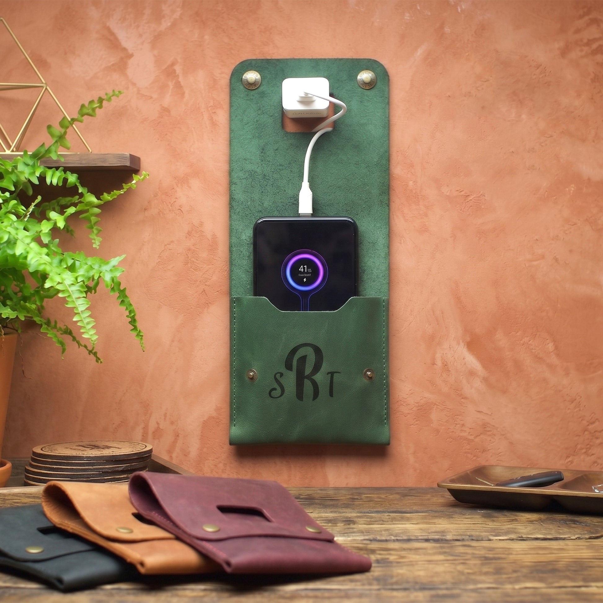 A green leather wall-mounted charging stand with a front pocket holding a smartphone, shown in a styled interior setting against a terracotta wall. Two additional charging stands in black and brown are visible on a wooden table in the foreground.