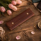A brown leather wallet or card holder with two gold-tone snap buttons, displayed on a rustic wooden table surrounded by pink tulips, rose petals, and small gold jewelry props.