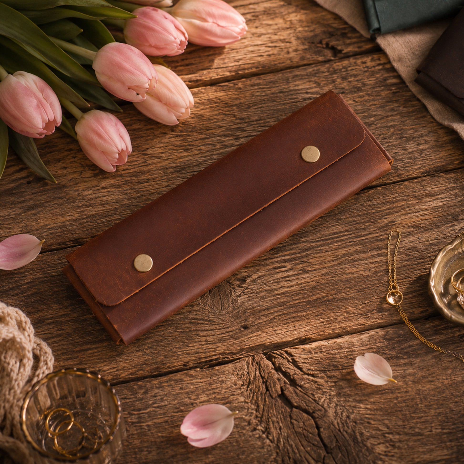 A brown leather wallet or card holder with two gold-tone snap buttons, displayed on a rustic wooden table surrounded by pink tulips, rose petals, and small gold jewelry props.