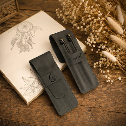 Two dark gray leather pen sleeves on a rustic wooden table, styled with an open sketchbook, dried flowers, and a wooden box. One sleeve is personalized with the name 'David' and the other features an engraved monogram 'G'.