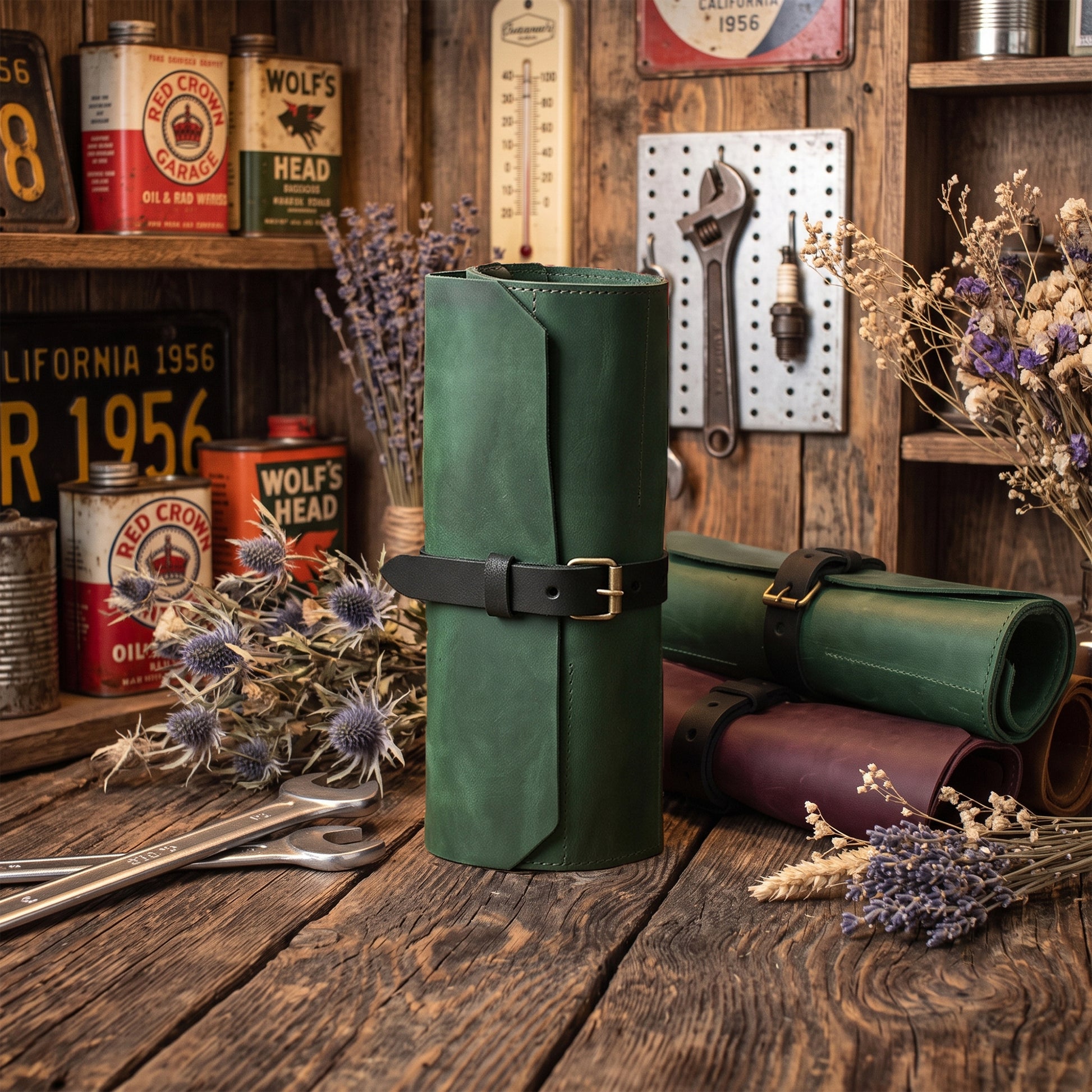 A dark green leather roll-up tool organizer with a black strap and brass buckle, displayed on a rustic wooden table alongside vintage oil cans, tools, and dried flowers.