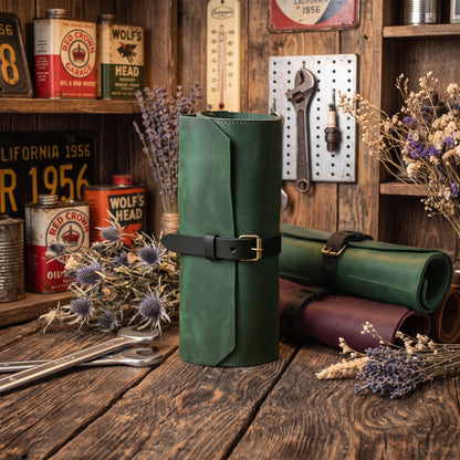 A dark green leather roll-up tool organizer with a black strap and brass buckle, displayed on a rustic wooden table alongside vintage oil cans, tools, and dried flowers.