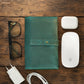 Green leather cable organizer, glasses, white Apple mouse, and charger on a wooden surface