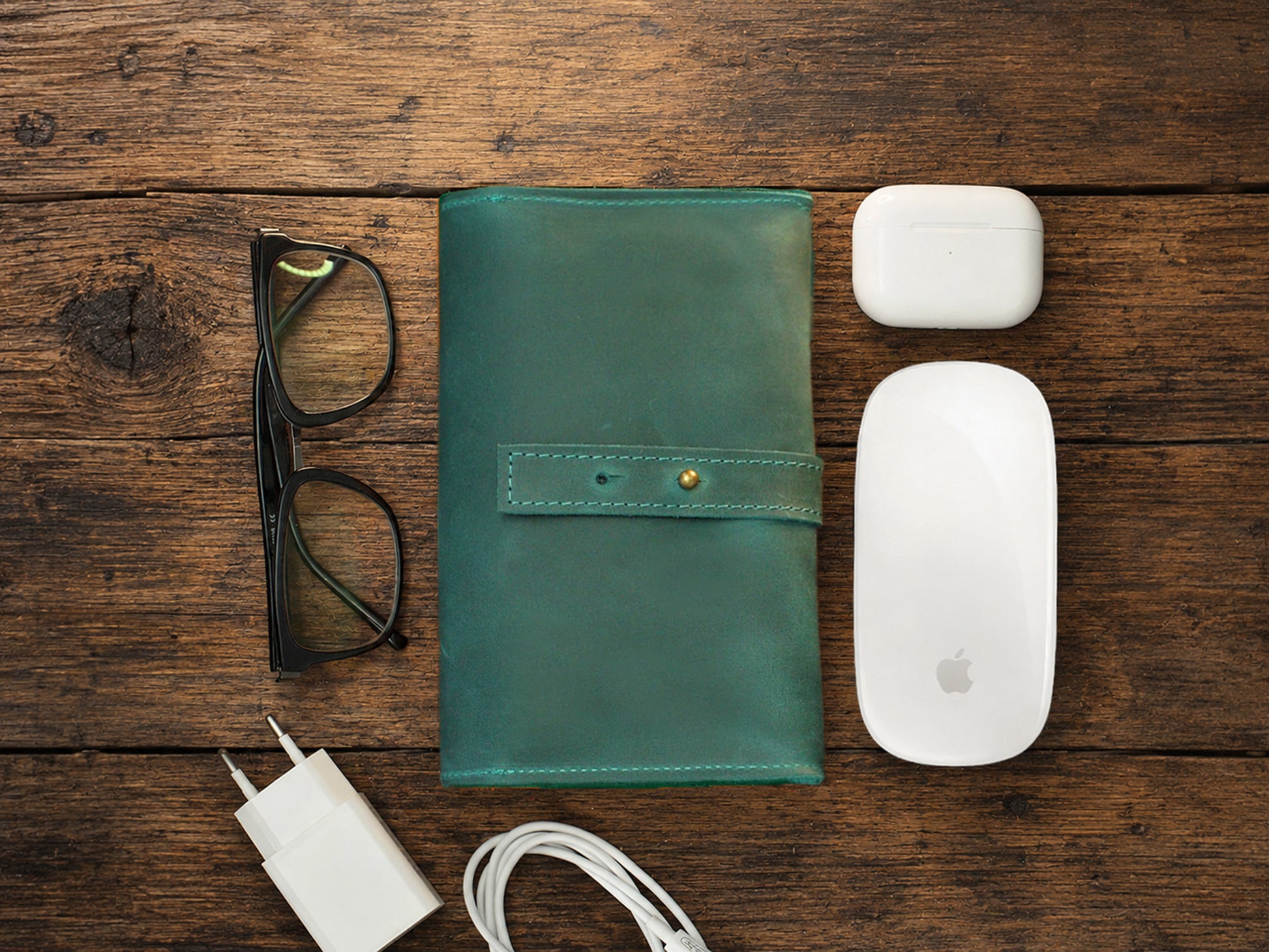 Green leather cable organizer, glasses, white Apple mouse, and charger on a wooden surface