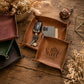 A brown leather square organizer tray on a rustic wooden table, holding a black car key fob and a smartphone. Another tray in front is engraved with 'Come Sweet Home'.