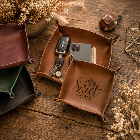 A brown leather square organizer tray on a rustic wooden table, holding a black car key fob and a smartphone. Another tray in front is engraved with 'Come Sweet Home'.