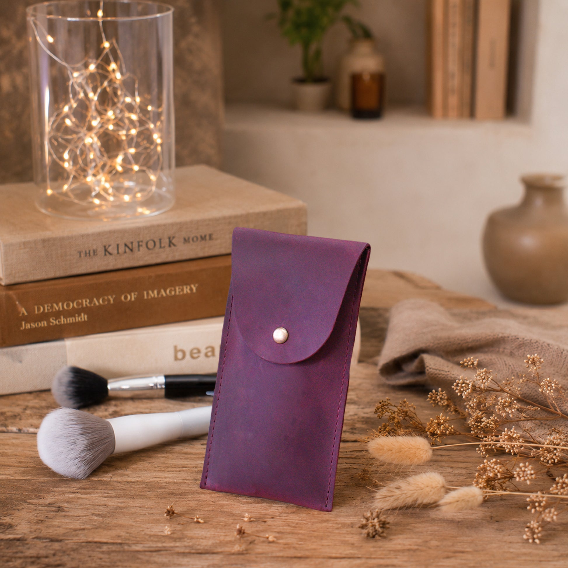 A purple leatherette pouch with a silver snap button, standing on a wooden table surrounded by makeup brushes, books, fairy lights, and dried flowers.