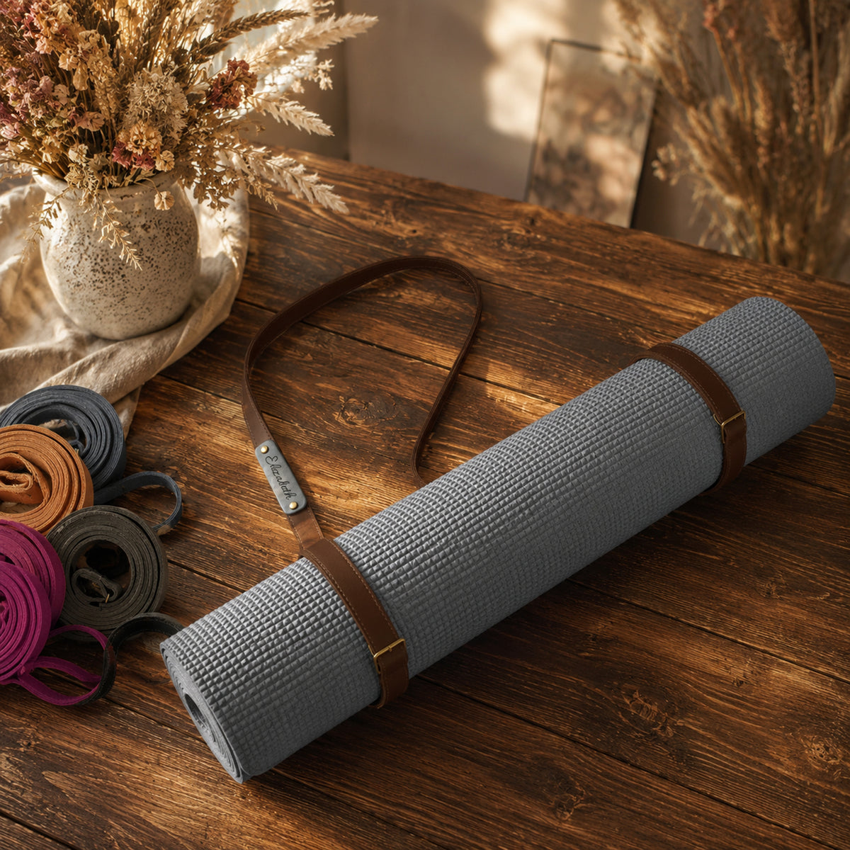 A rolled gray textured yoga mat with brown straps and a matching strap laid beside it on a rustic wooden table, with dried flowers and other rolled mats in the background.