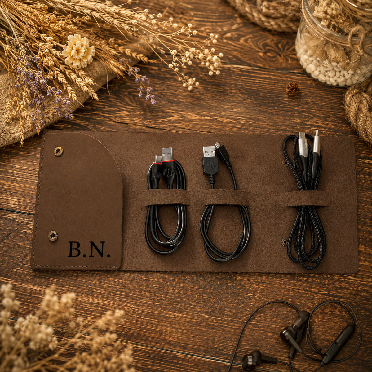 A brown leather cable storage roll laid on a wooden surface, holding three coiled black cables. The roll has brass grommets and the initials 'B.