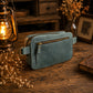 A dusty blue leather crossbody pouch bag with gold-toned hardware, resting on a rustic wooden table surrounded by dried flowers, a vintage lantern, and a book.