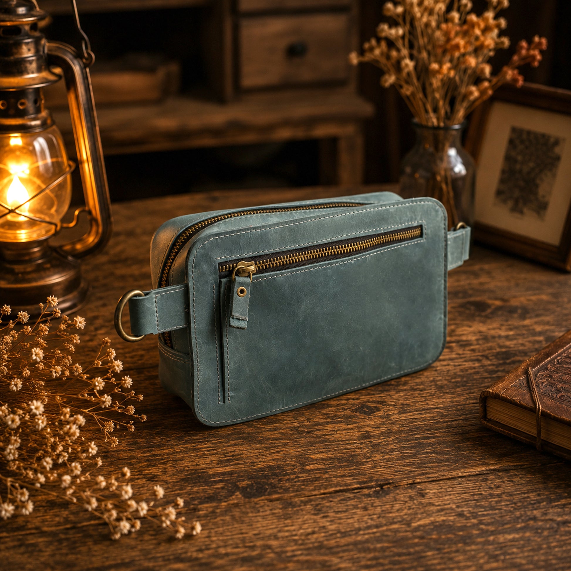 A dusty blue leather crossbody pouch bag with gold-toned hardware, resting on a rustic wooden table surrounded by dried flowers, a vintage lantern, and a book.