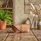 A tan leather pouch or small case with a wrap strap and metal snap, placed on a rustic wooden table in a styled interior setting with a potted fern, dried flowers, and a woven wall hanging in the background.