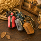 Four small zippered pouches or coin purses in coral pink, gray, dark teal, and tan are arranged on a wooden surface. The tan pouch is embroidered with the name 'BENTLEY' and the gray pouch is embroidered with 'Lola'.