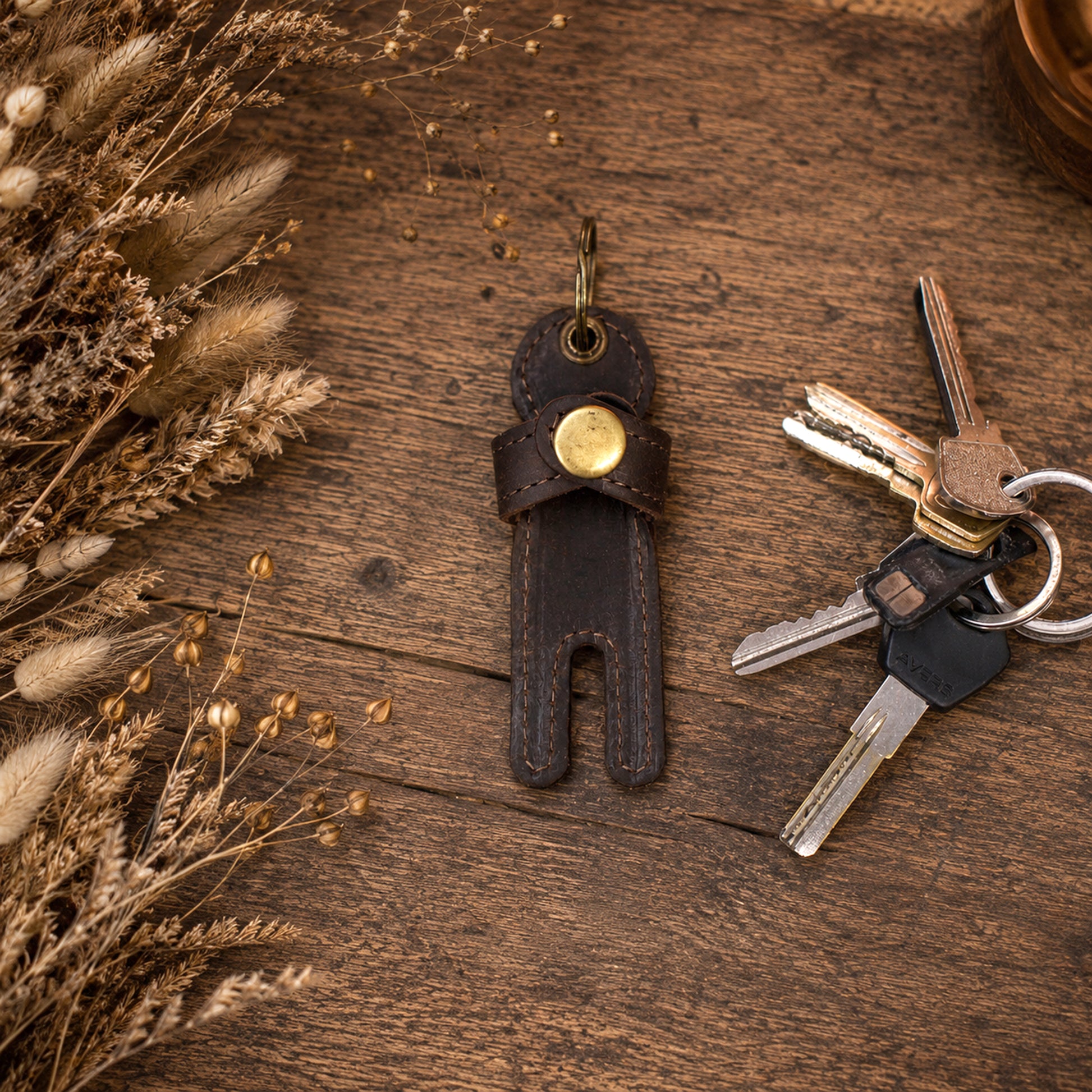 A dark brown leather key holder keychain with a gold-tone snap button, shown with keys on a rustic wooden surface next to dried decorative grasses.