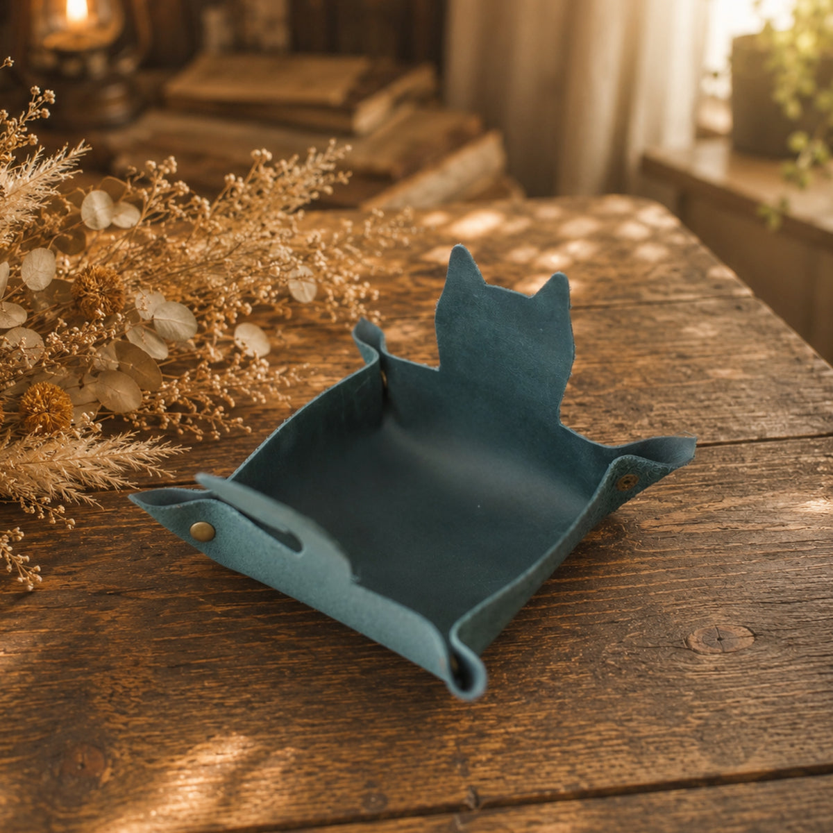 A teal blue felt cat-shaped tray with a small metal snap, resting on a rustic wooden table next to dried flowers.