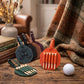 Three leather golf tee holders in dark teal, brown, and forest green are arranged on a rustic wooden table with golf tees inserted. A white golf ball sits nearby, with books, a vase of dried flowers, and a plaid blanket in the background.