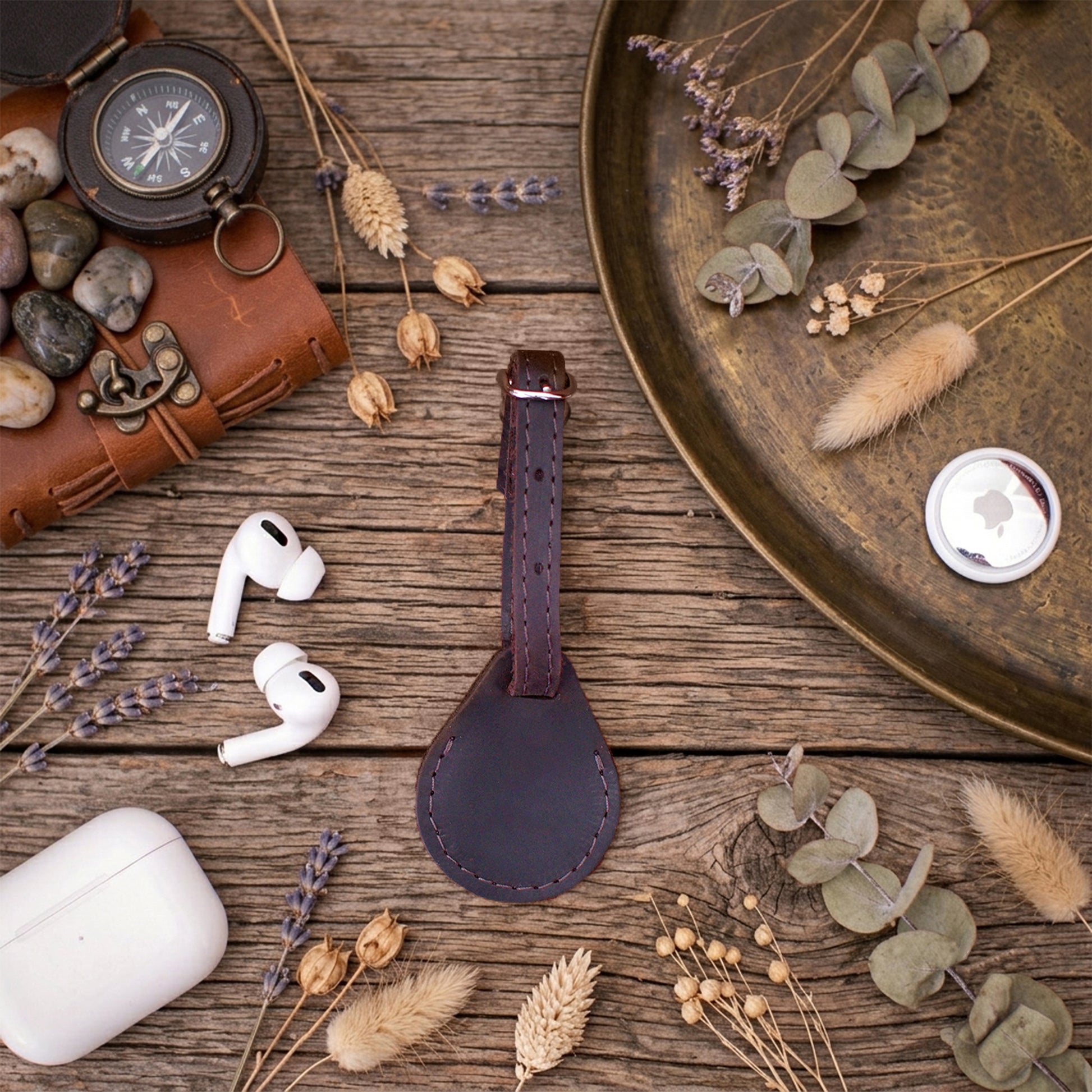 A dark brown leather luggage tag with a stitched loop, styled in a flat-lay arrangement on a rustic wooden table with a compass, AirPods, a leather case, and dried botanicals.