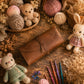 A brown leather crochet hook organizer case lies on a rustic wooden surface, surrounded by yarn balls, crocheted teddy bears, crochet hooks, and dried floral decorations.