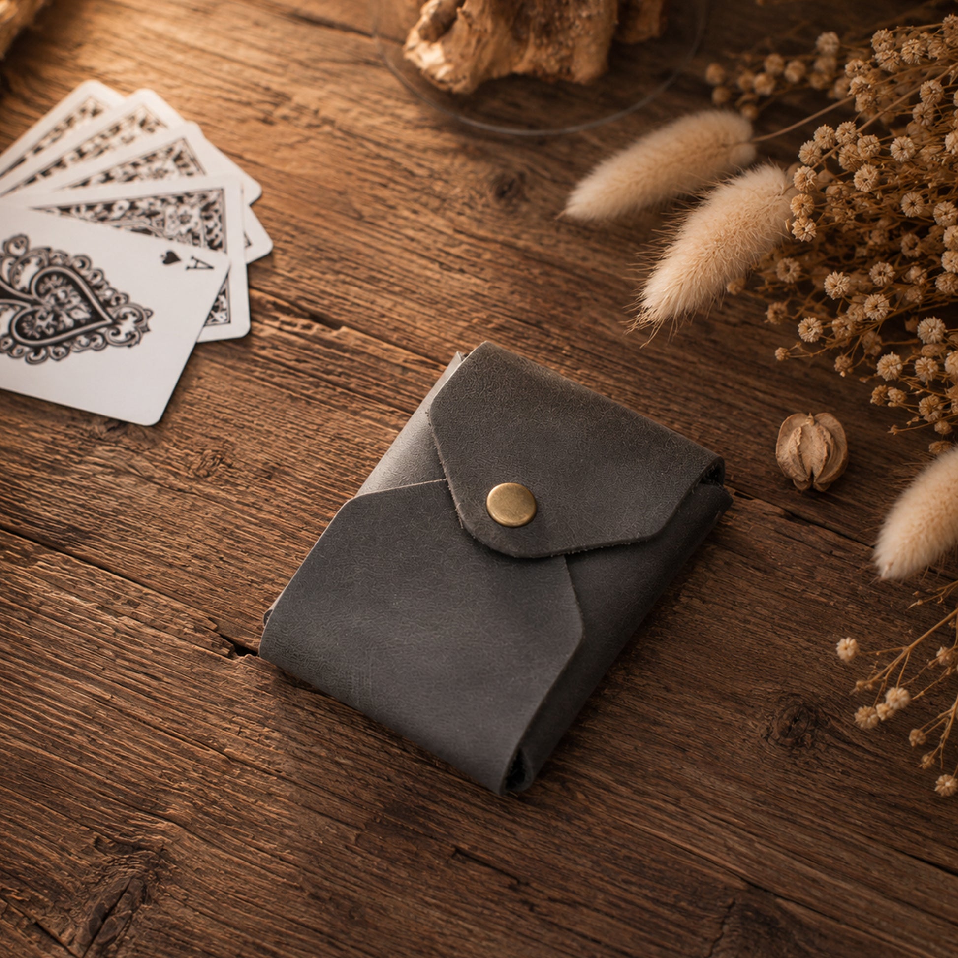 A dark gray leather card holder wallet with a gold snap button, resting on a rustic wooden table surrounded by dried flowers, playing cards, and a wooden bead.