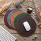 Four round leather mouse pads in dark brown, green, orange-brown, and burgundy, arranged in a fan shape on a rustic wooden desk with a white computer mouse on the front pad, styled with dried grasses, a keyboard corner, and earbuds.