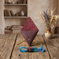 A diamond-shaped burgundy leather stand with brass hardware, displayed on a rustic wooden table with blue earphones and a small vase of dried lavender and wheat in the background.