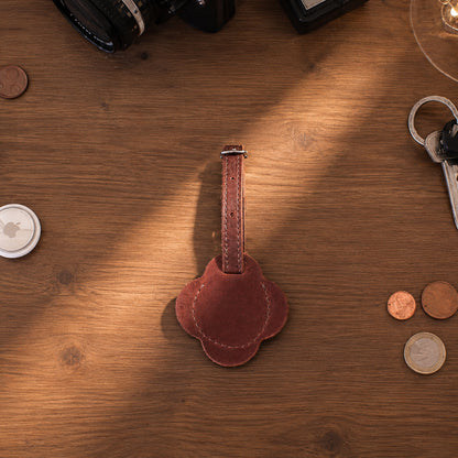 A brown leather keychain or luggage tag with a decorative scalloped end, placed on a wooden tabletop surrounded by everyday objects like coins, keys, a camera, and a glass.