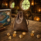 A brown leather drawstring bag stands on a wooden table, surrounded by gold-toned polyhedral dice. A fantasy map box and books are visible in a cozy, candle-lit medieval-style room with stone windows and a fireplace.