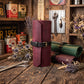A leather roll-up sleeve or organizer standing upright on a rustic wooden table, with matching rolled leather items and dried flowers around it. The main product is a burgundy leather sleeve with a black strap and gold-tone buckle.
