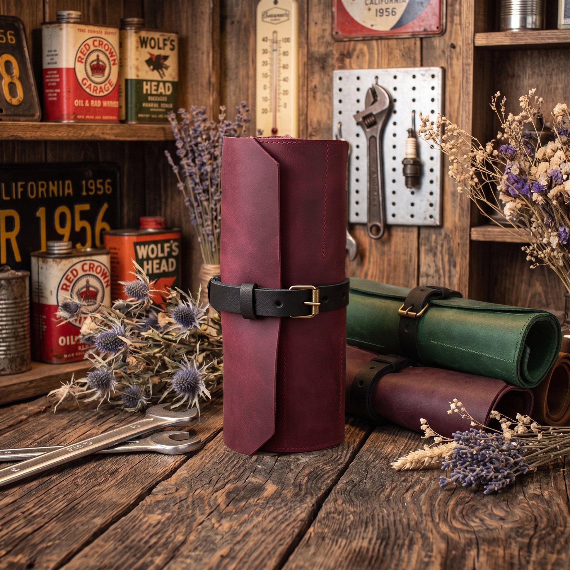 A leather roll-up sleeve or organizer standing upright on a rustic wooden table, with matching rolled leather items and dried flowers around it. The main product is a burgundy leather sleeve with a black strap and gold-tone buckle.