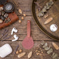 A dusty rose leather luggage tag with a stitched loop handle, styled in a flat-lay arrangement on a rustic wooden table with eucalyptus leaves, dried grasses, a compass, a brown leather case, white AirPods, and a white charging case.