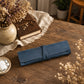 A rolled dark blue leather journal or notebook cover tied with a matching strap, placed on a rustic wooden table with dried flowers, a book, and a small dish in a styled indoor setting.