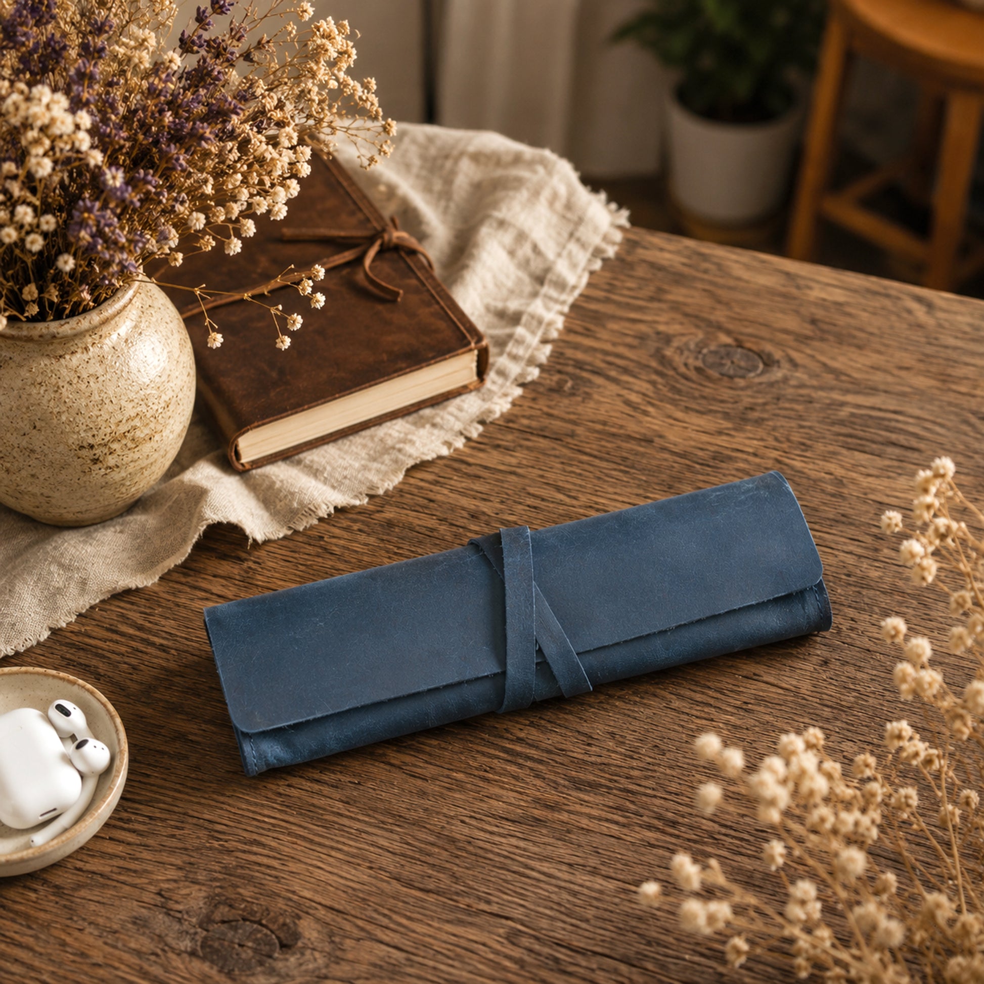 A rolled dark blue leather journal or notebook cover tied with a matching strap, placed on a rustic wooden table with dried flowers, a book, and a small dish in a styled indoor setting.