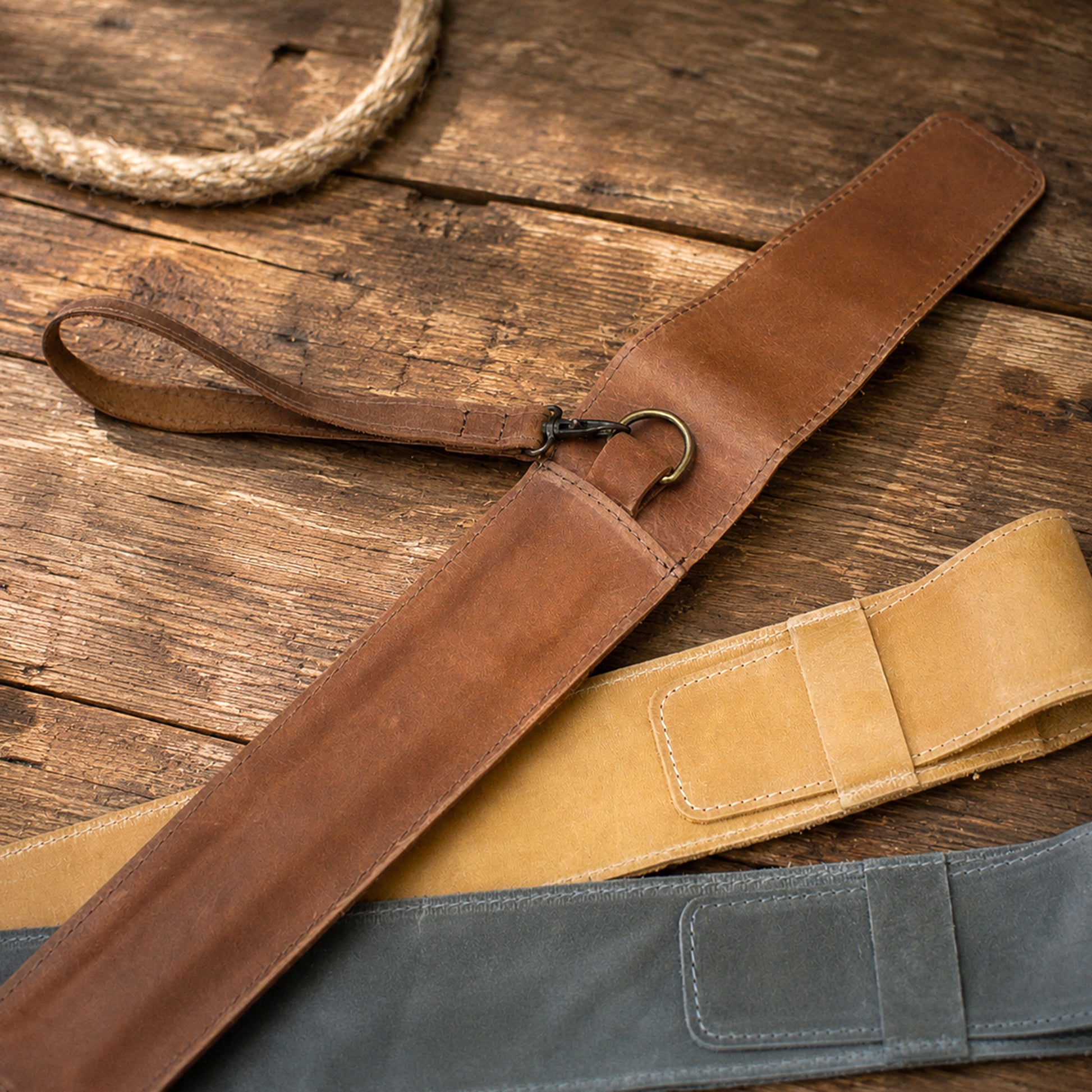 Three leather belt-like straps laid diagonally on a rustic wooden surface: a dark brown strap with a loop and metal clip, a tan strap, and a muted blue-gray strap. A rope is partially visible in the upper left.