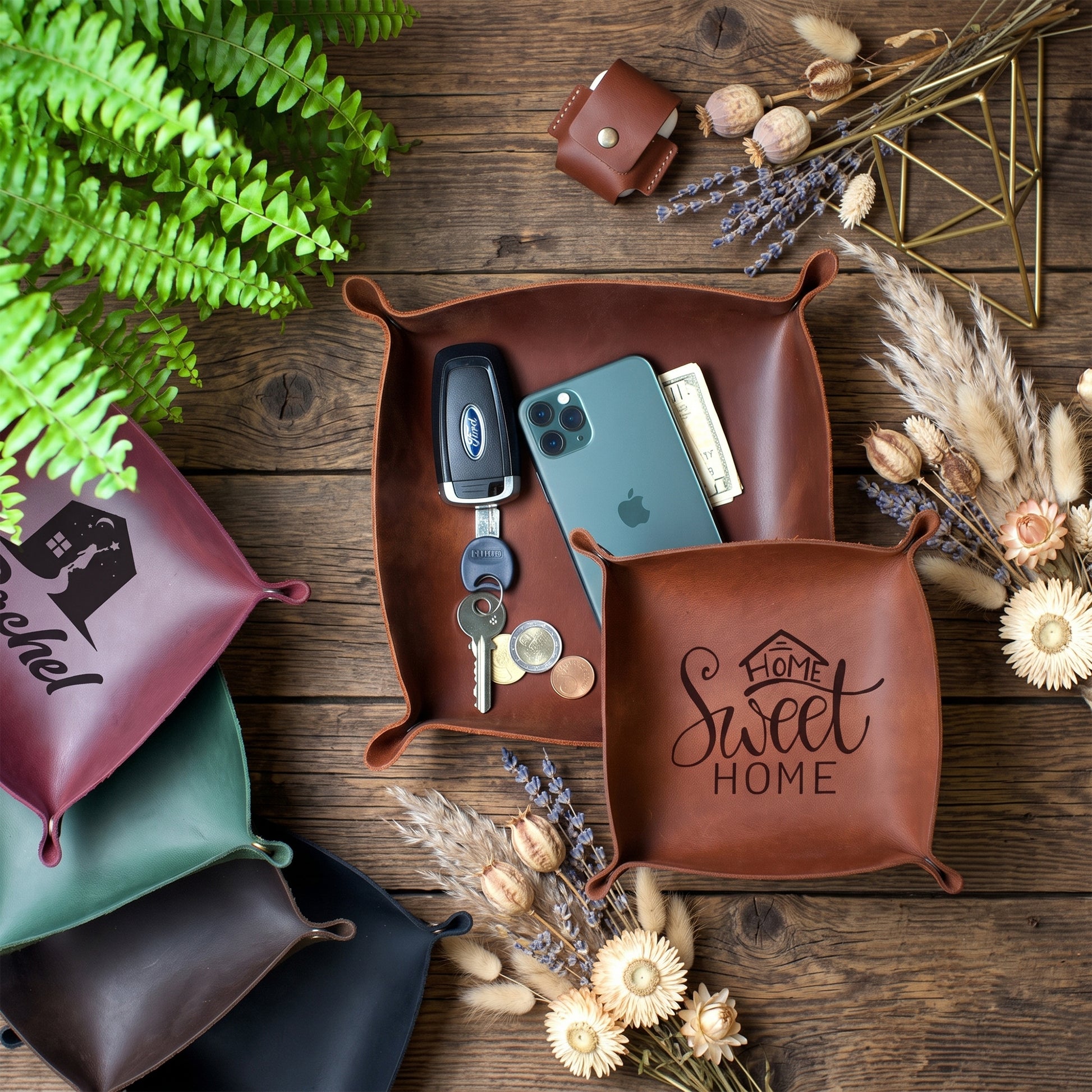 A styled flat-lay of leather square pouches on a rustic wooden surface. The main focus is a medium brown leather pouch in the center containing a car key fob, a green smartphone, and cash, with the front printed with 'home Sweet HOME'.