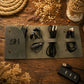 A dark olive green roll-up organizer pouch laid on a rustic wooden surface, holding a black power adapter, earbuds, two black cables, and a black multi-port USB charger. The pouch has a monogrammed 'H' on the left flap and is styled with dried flowers and rope around it.