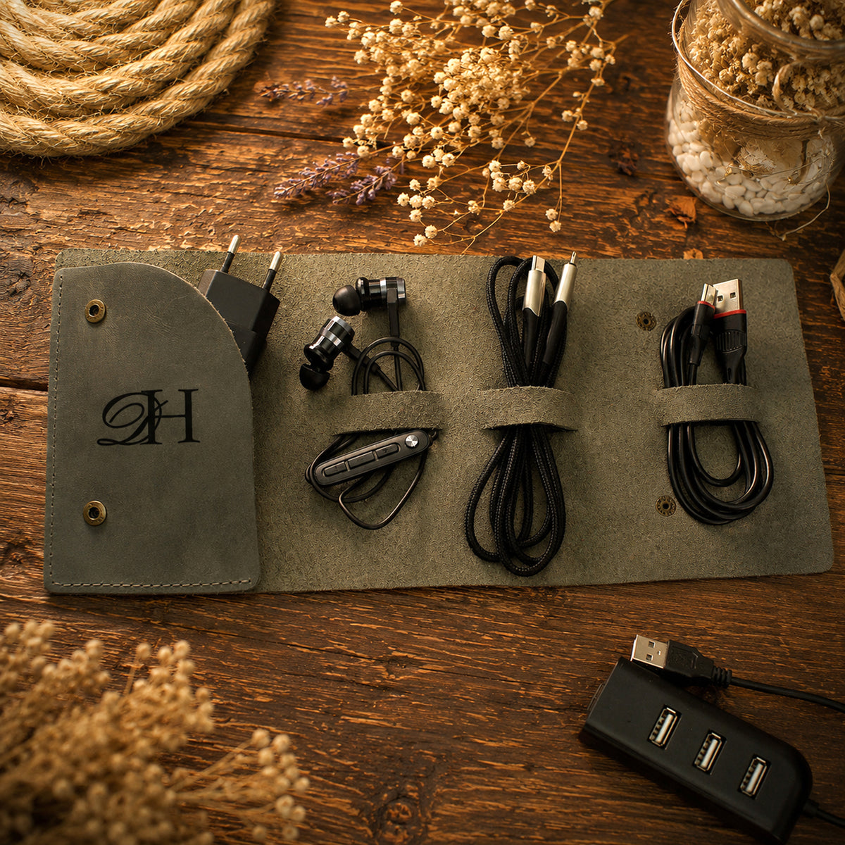 A dark olive green roll-up organizer pouch laid on a rustic wooden surface, holding a black power adapter, earbuds, two black cables, and a black multi-port USB charger. The pouch has a monogrammed 'H' on the left flap and is styled with dried flowers and rope around it.