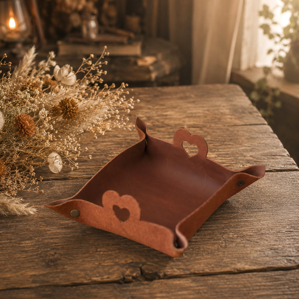 A brown leather tray with heart-shaped cutouts on the handles and base, displayed on a rustic wooden table next to dried flowers.