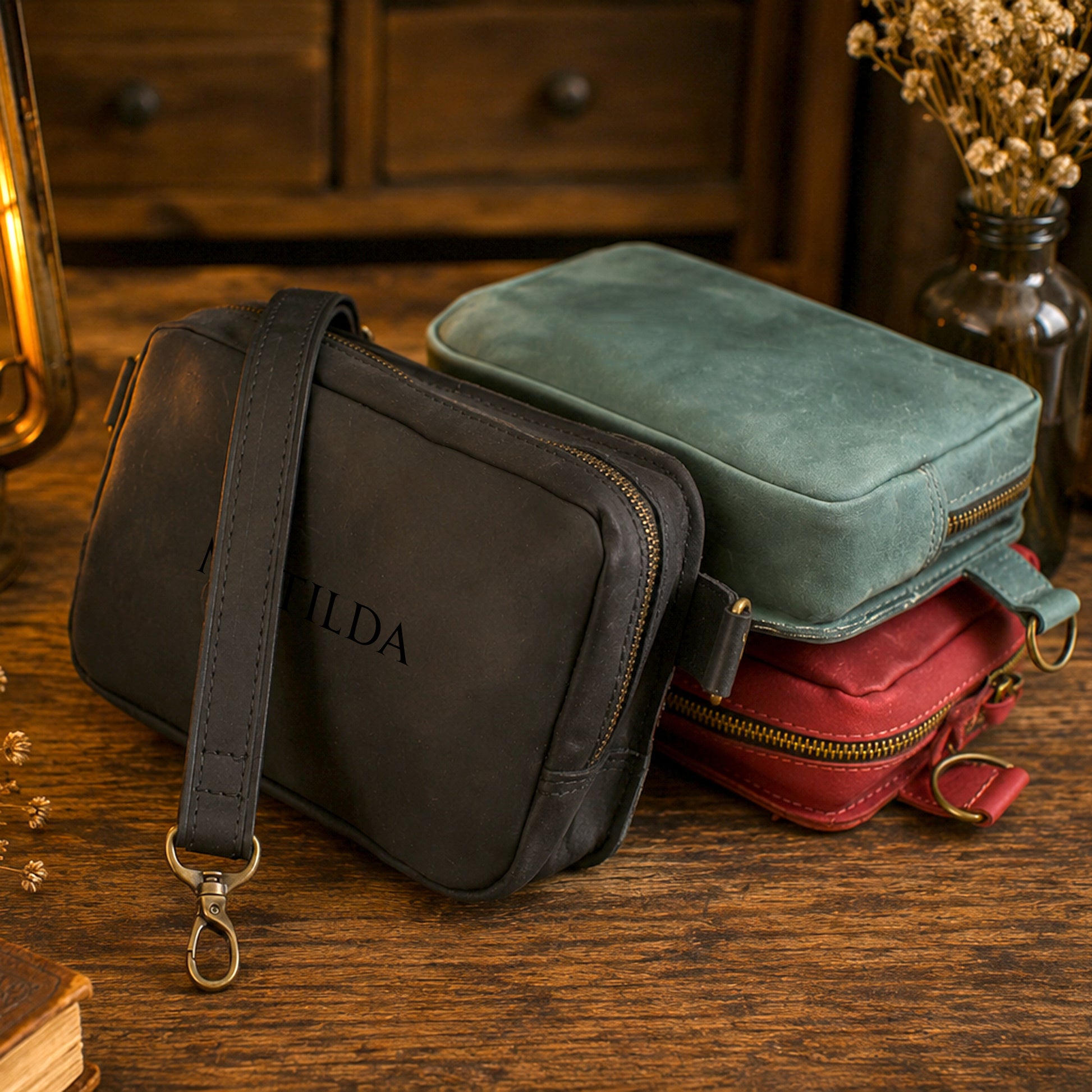 Three leather crossbody pouches in black, sage green, and red are stacked on a rustic wooden table. The black pouch in the foreground features an embossed 'ILDA' logo and a gold-tone clasp, with a wooden cabinet and a glass vase of dried flowers softly blurred in the background.