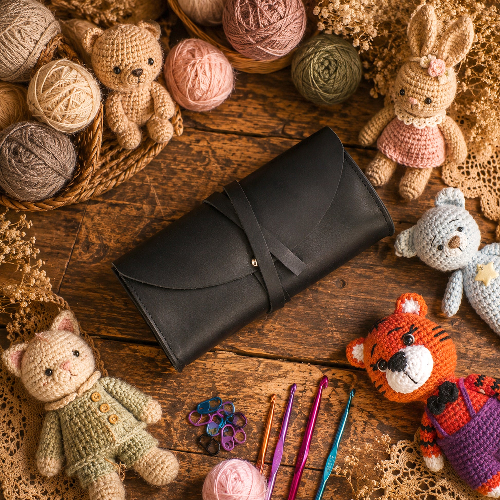 A black leather zippered pouch or case with a strap closure, placed on a rustic wooden surface surrounded by crocheted toys, yarn balls, crochet hooks, and lace decorations.