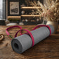 A gray textured yoga mat rolled up and secured with a bright pink strap, placed on a rustic wooden table in a warm, softly blurred home interior setting.