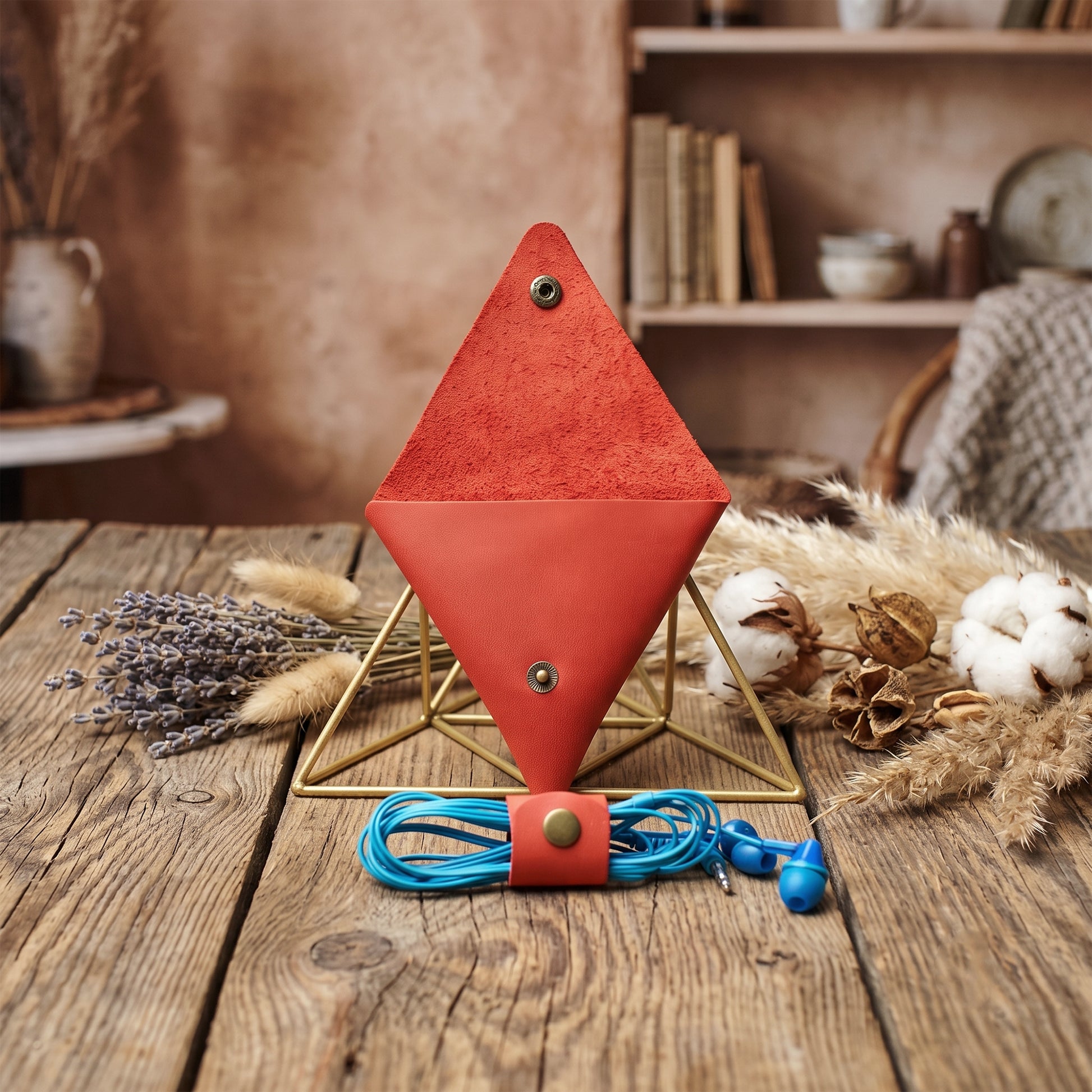 A red diamond-shaped leather organizer stand on a gold metal frame, holding a blue coiled cord with a matching red snap closure. The setup is styled on a rustic wooden table with dried lavender, cotton bolls, and books in the background.