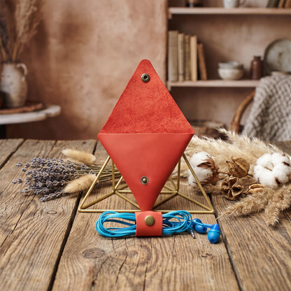 A red diamond-shaped leather organizer stand on a gold metal frame, holding a blue coiled cord with a matching red snap closure. The setup is styled on a rustic wooden table with dried lavender, cotton bolls, and books in the background.
